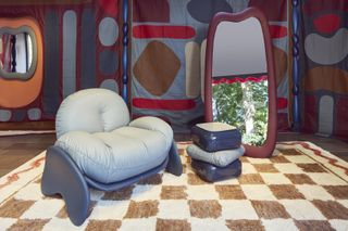 A design showroom presents whimsical furniture including a light blue upholstered armchair, a "squashed" coffee table in the same tone, a monster-like mirror, and vibrantly colored tapestry covering both walls and floors.