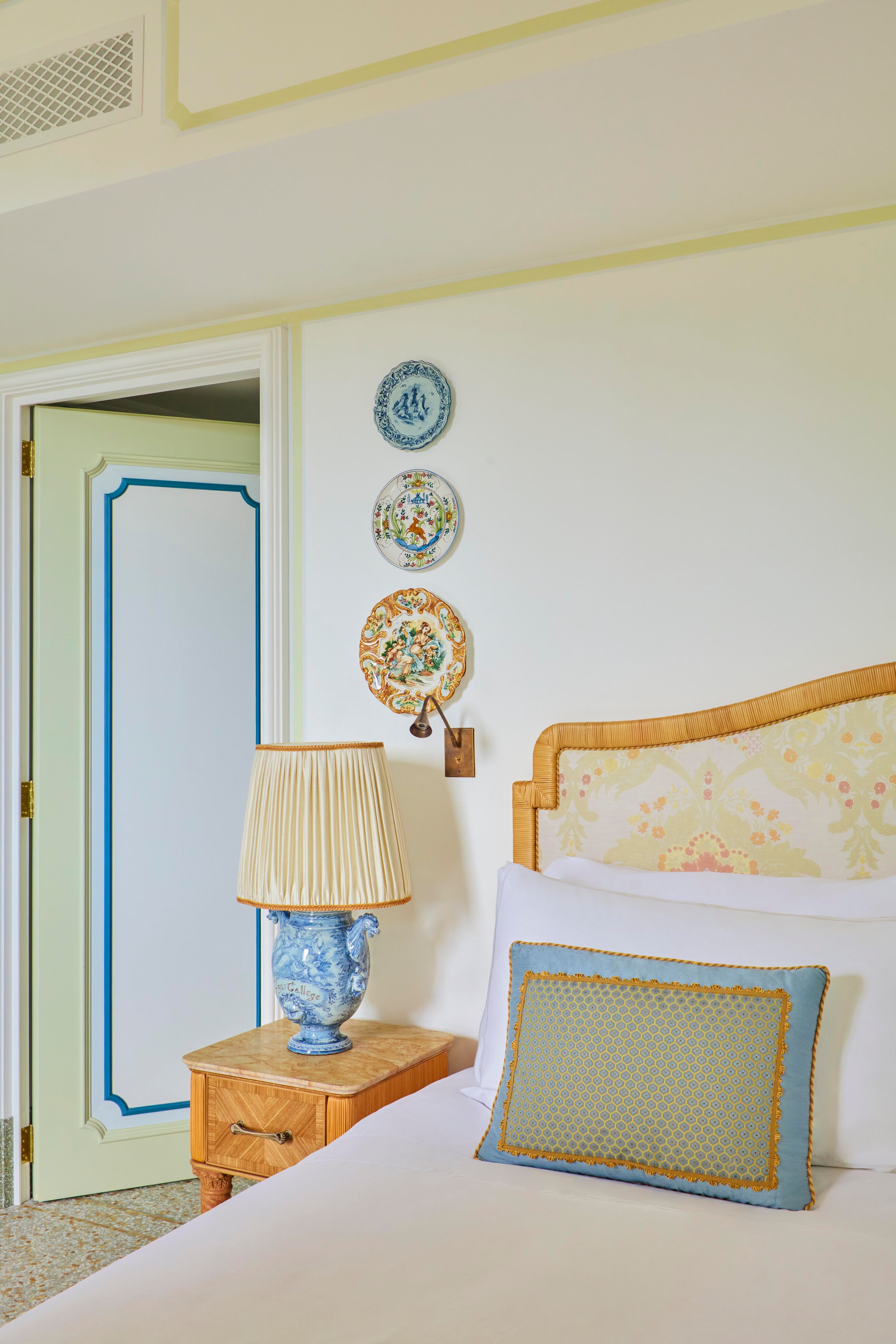 A pale wooden bed with an artisanal wooden bedside table, blue ceramic lamp and three decorative plates hung on the wall