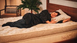 Our mattress tester Lauren Jeffries lies on the Birt Elite Natural Mattress in a side sleeping position. The mattress is on a wooden bed frame in a decorated room. She uses a high loft pillow to cushion her head, neck and shoulders