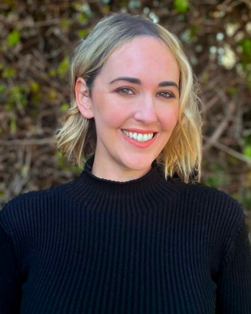 a headshot photo of therapist and councelor Cheryl Groskopf: a lady in a black long sleeve standing outside in front of a hedge with blond tied up hair smiling at the camera