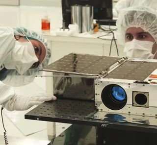 two scientists in bunny suits stare at a cubesat on a table.