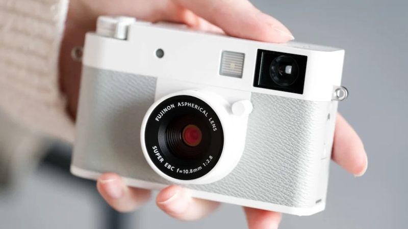 A person holds a stylish, white Fujifilm X Half camera featuring an aspherical Fujinon lens, with a soft-focus background