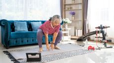 woman exercising at home in front of an ipad, she's mid squat and touching the floor. there's a blue sofa behind her and some exercise equipment to the side 