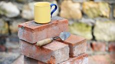 A pile of bricks outside with a cement trowel and a yellow mug with a blue handle 