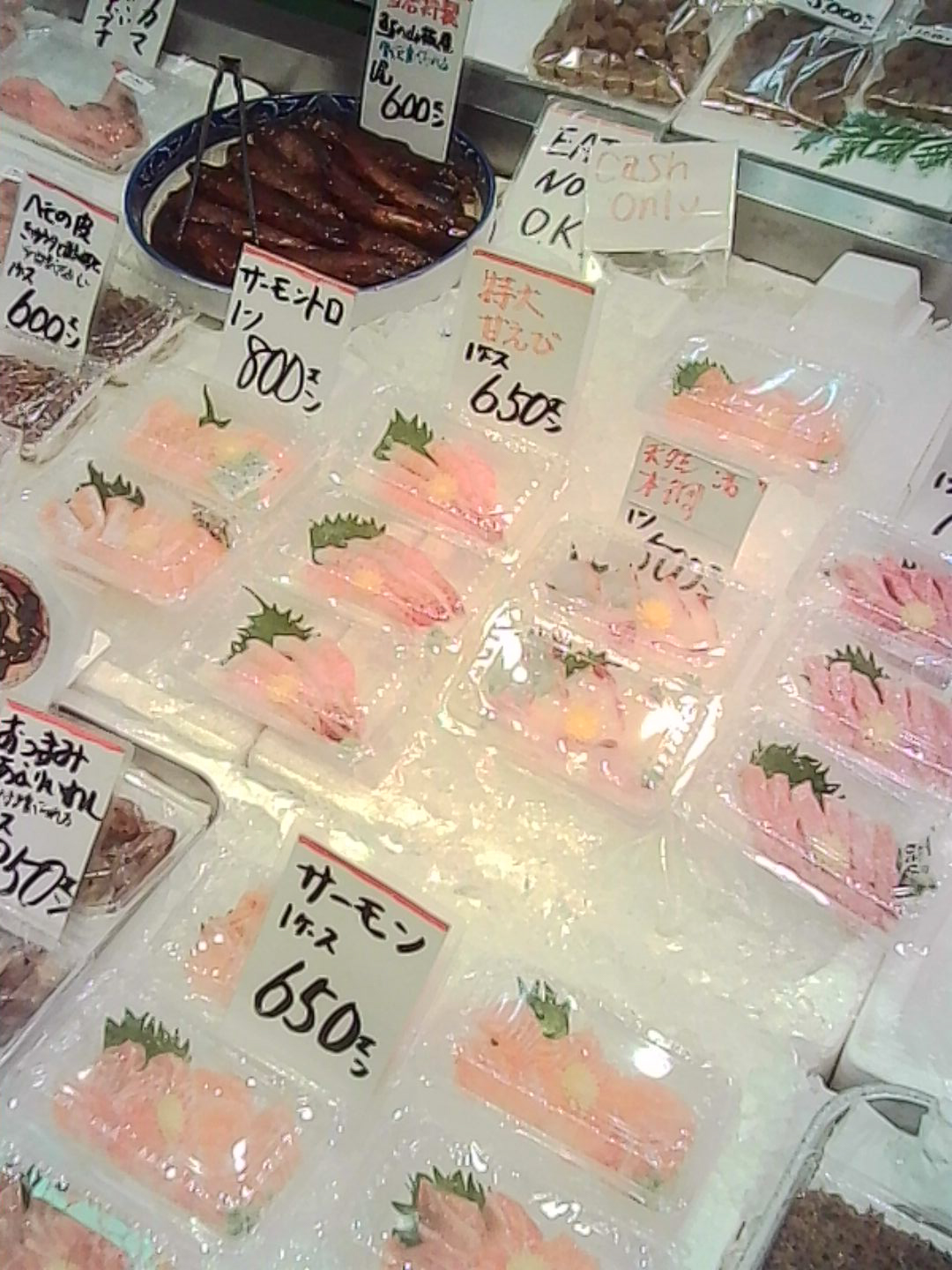 Packaged seafood displayed on ice with handwritten price signs in a Japanese market.