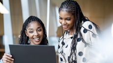 Two women smile as they look at a laptop together in an office.
