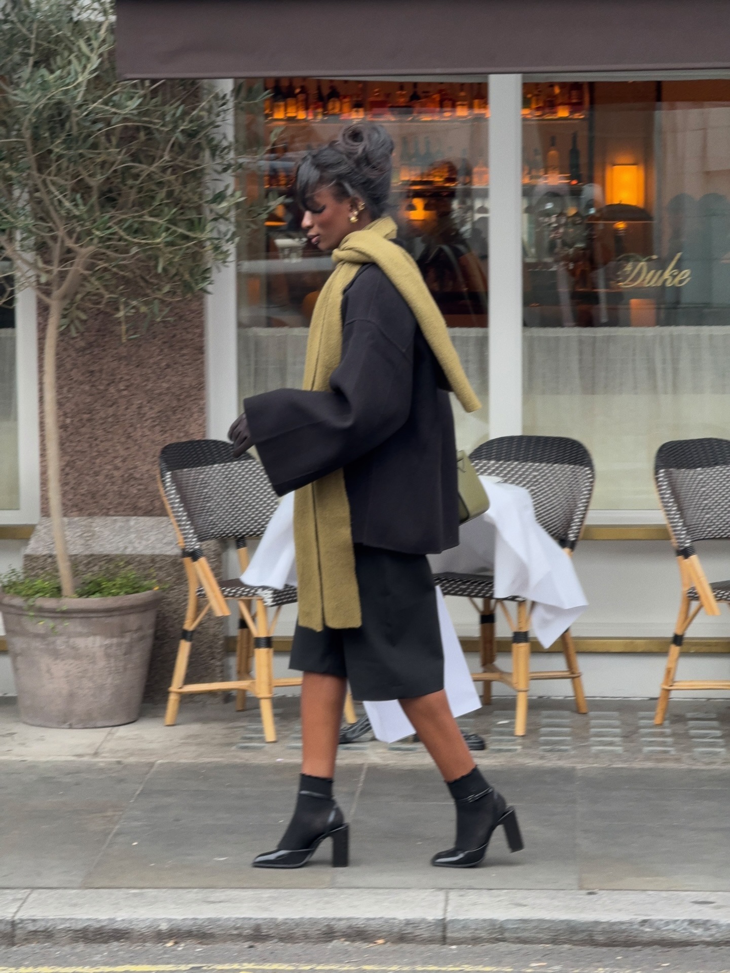 Fashion person Liv Madeline shows what to wear in February. She wears black bermuda shorts, a black Wrap coat and square toe black heel with black socks. She adds some colour with over green accessories, namely a scarf and bag. This image was taken recently from her Instagram.
