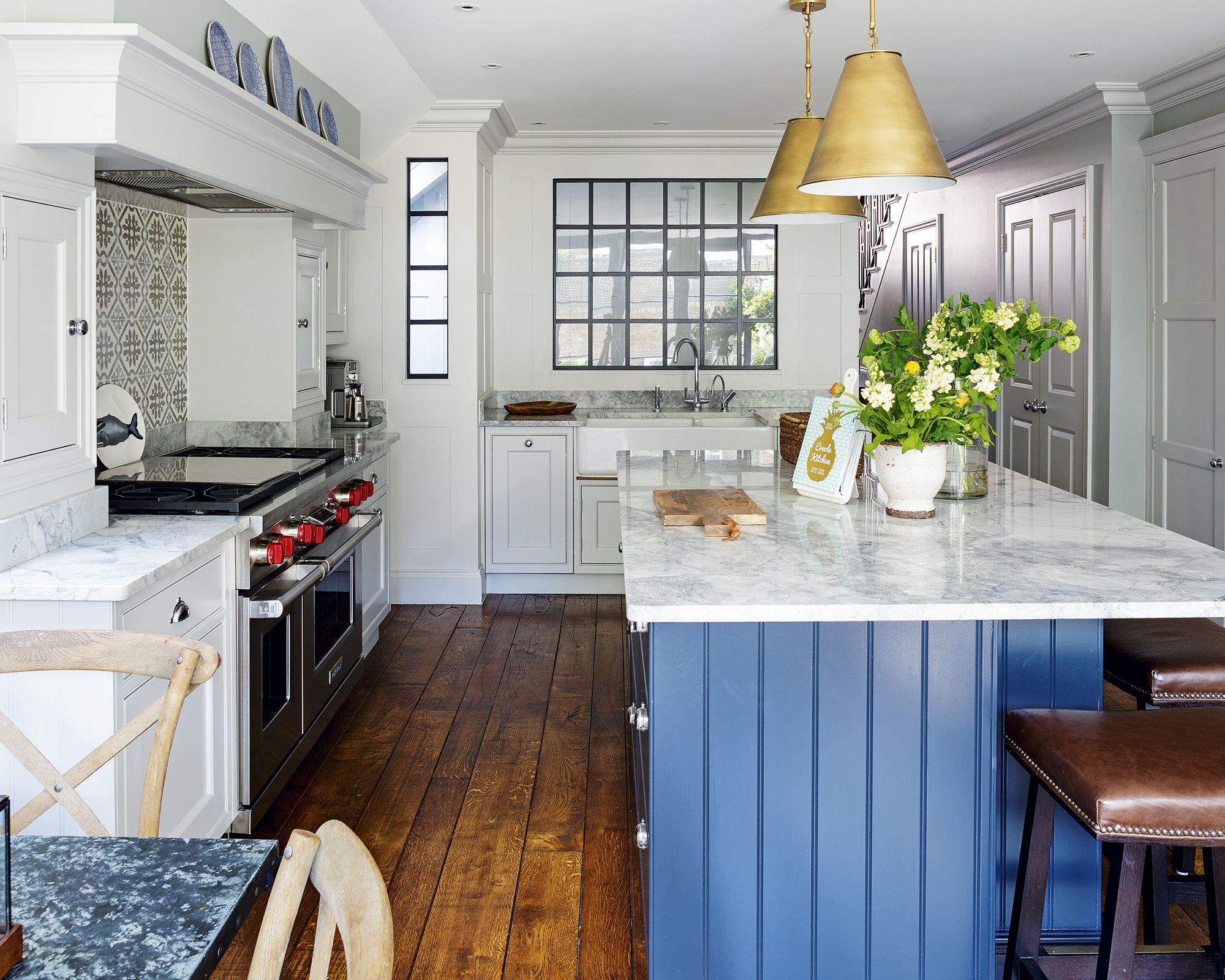An example of small kitchen ideas showing a kitchen with cream cabinets and marble worktops and a blue, wood paneled island below two gold pendant lamps