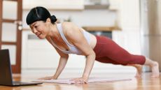 A woman in a tank top and leggings performs push-ups on a yoga mat with a laptop screen open in front of her.