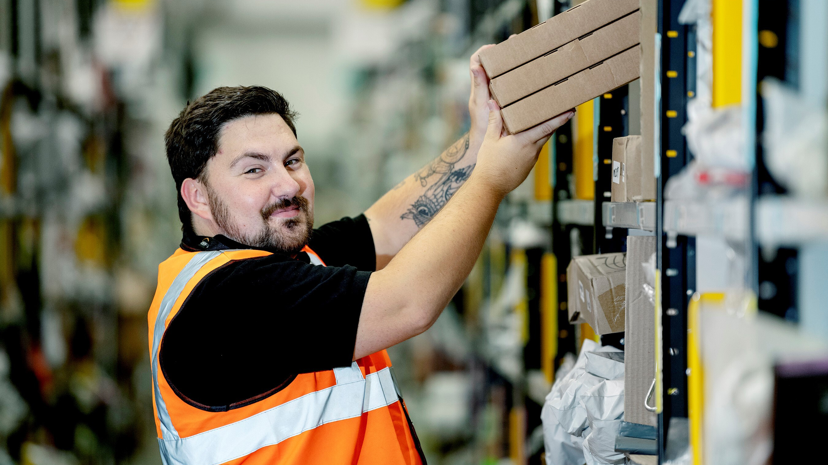 A man working in a warehouse lifts a stack of cardboard boxes from a shelf. He wears an orange high-visibility vest over a black shirt, has short dark hair, a beard, and a visible tattoo on his right arm.