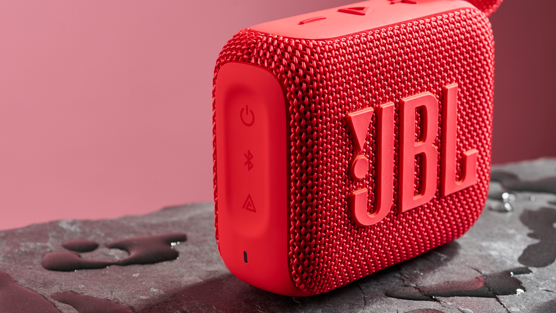 The side panel and control buttons of a red JBL Go 4 pictured against a pink background. It is sitting on a dark stone surface with puddles of water around it.