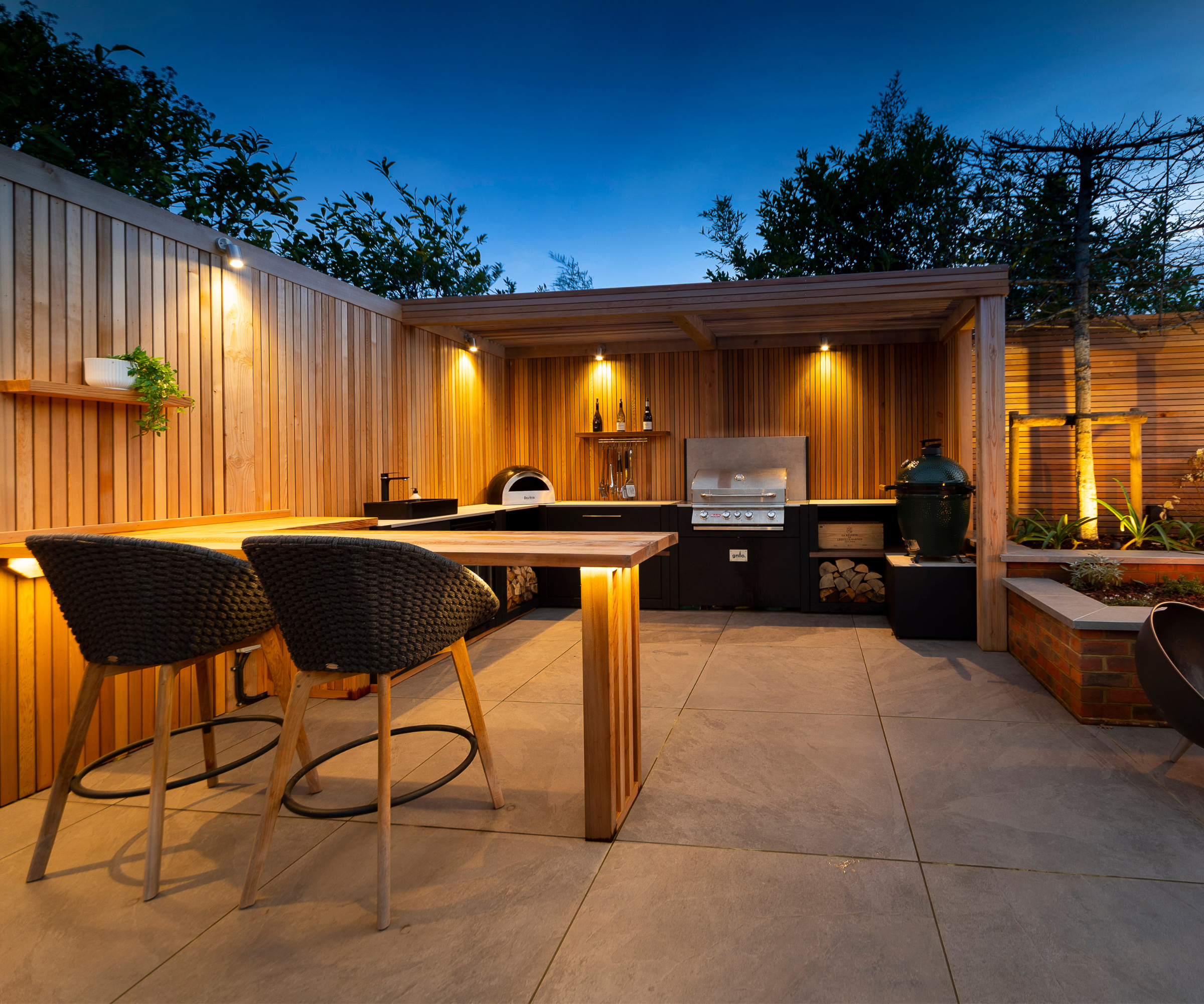Outdoor kitchen lit at night with wooden paneling along the fence that connects to the pergola covering the kitchen area