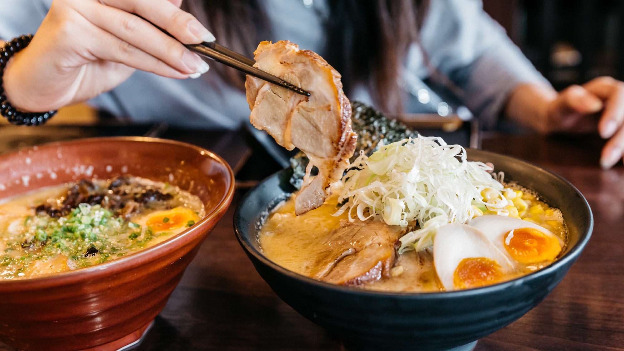 side view of two bowls of tonkotsu ramen, one has a woman picking up a piece of chashu with brown chopsticks