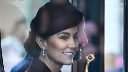 Kate Middleton wearing a maroon hat and coat with diamond earrings riding in a carriage next to Prince William