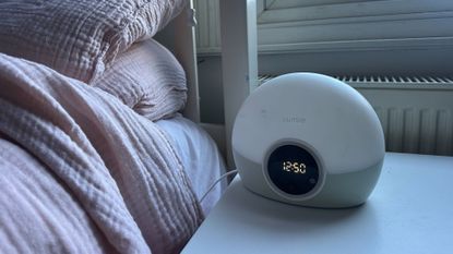 A Lumie alarm clock sits on a bedside table next to the corner of a bed.