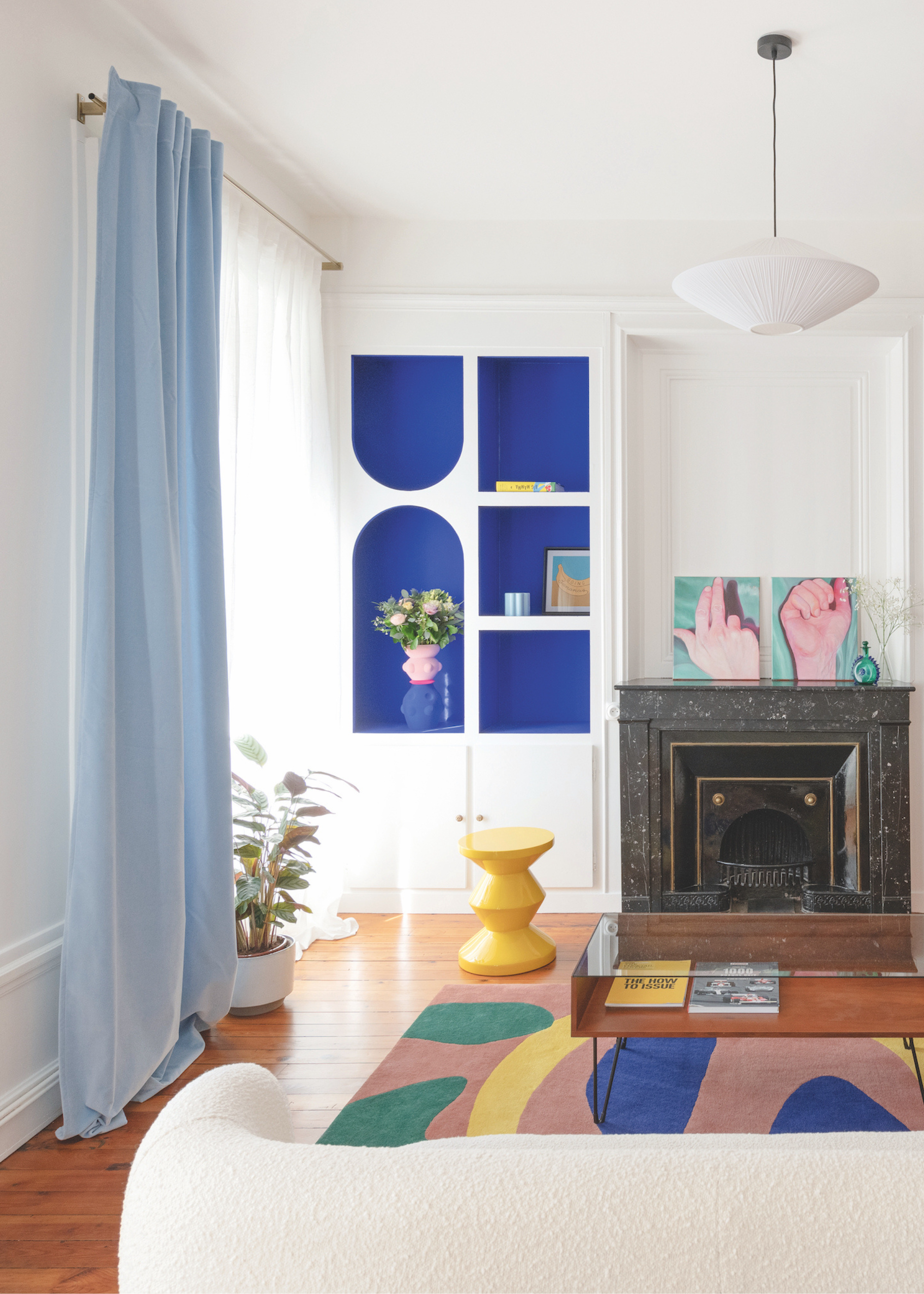 a modern living space with white walls and bold contrasting colors in geometric cut out shapes with pale blue floor length curtains, a black fireplace, a cream sofa, cobalt blue alcove shelving, and a modern yellow stool