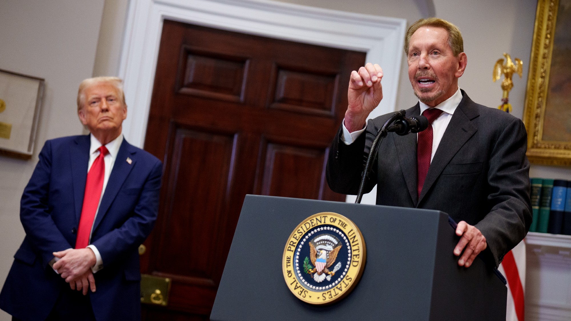 Larry Ellison speaks at a podium in the Roosevelt Room of the White House, while Donald Trump looks on