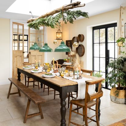 dining room with patio doors with long wooden dining table, antique school chairs and bench with a branch hanging overhead covered in greenery with three green pendant lights