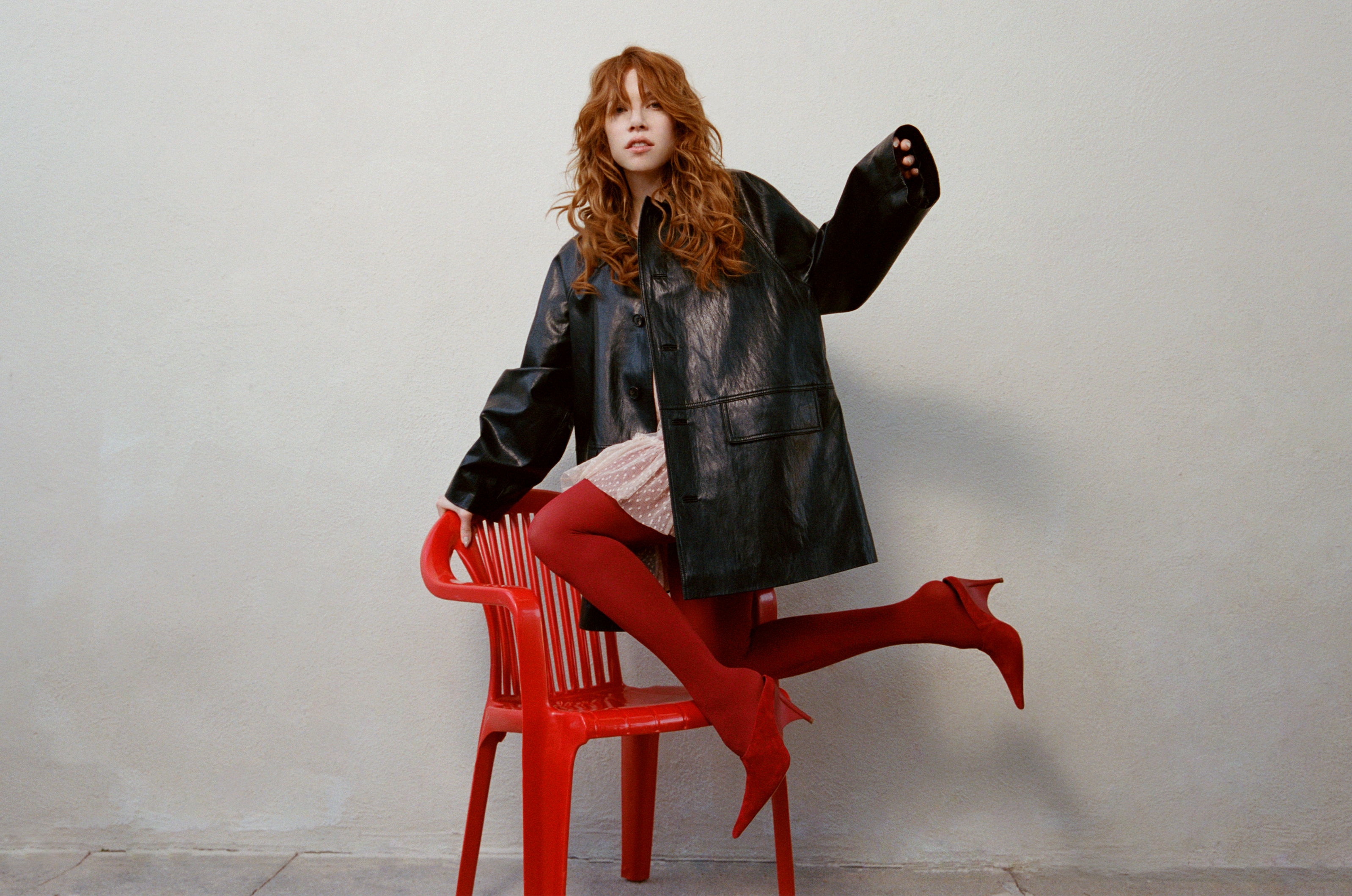 carly rae jepsen poses climbing onto a red chair wearing red tights and an oversized leather jacket