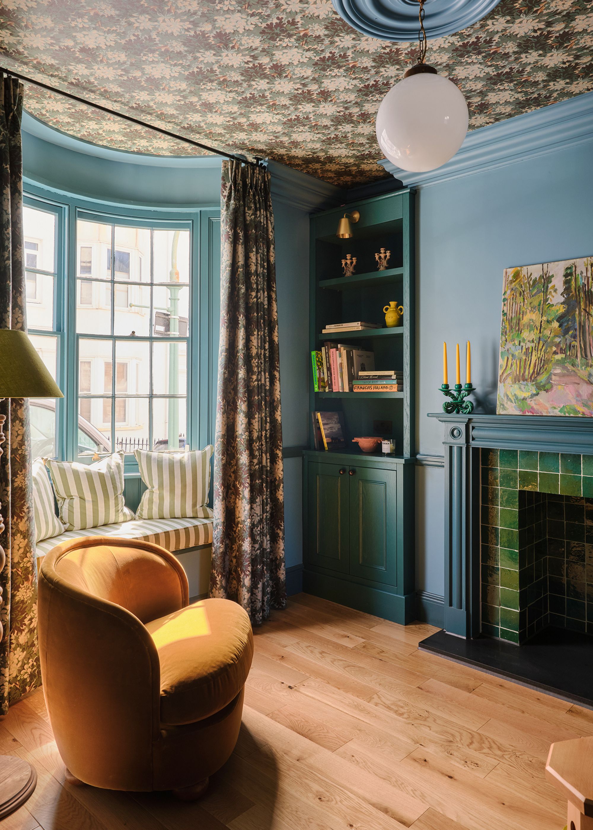 A living room with blue walls and a bay window, floral wallpaper on the ceiling, and floral curtains. Wooden flooring with a teal fireplace and alcove shelving, banquette window seating, and a modern ochre armchair.
