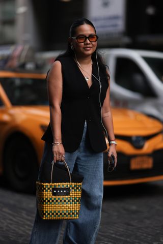 A Fashion Week guest is seen wearing black sunglasses with orange lenses, golden necklaces, a black waistcoat, several silver bracelets and rings, a silver watch, a square basket bag form MARNI with black/yellow/green leather, wide blue jeans and black leather boots with silber toe cap outside before the Naeem Khan Show on September 12, 2023 in New York City.