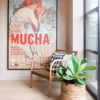Wicker armchair and houseplant next to an oversized art nouveau framed print on a plaster wall