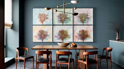 blue dining room with timber table and timber and leather chairs, six colorful paintings of coral on the wall, a sculptural pendant light, a vase with foliage, the table styled with a bowl and a bowl of oranges, and linen blinds over a bright window