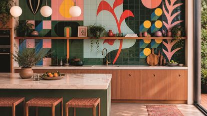 a modern kitchen with timber cabinetry, a green island with marble top and three rattan bar stools, and a colorful tiled mural backsplash on the back wall with a floating shelf in front