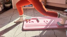 Bottom half of person exercising using a Pilates board, with one foot on a slider on the board and the foot behind the person’s front foot