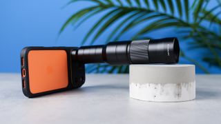 A photo of the Sandmarc 10x telephoto lens on an orange iPhone 17 Pro against a blue background