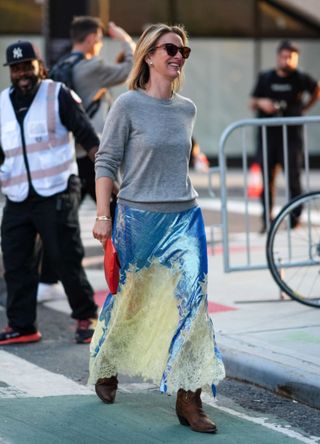 A woman wearing black sunglasses, silver earrings, dark gray sweater, gold bracelets and a shiny Tory Burch midi skirt.