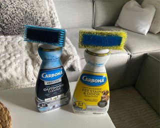Two bottles of Carbona upholstery cleaner, one yellow the other blue, on white wooden coffee table in front of grey sofa