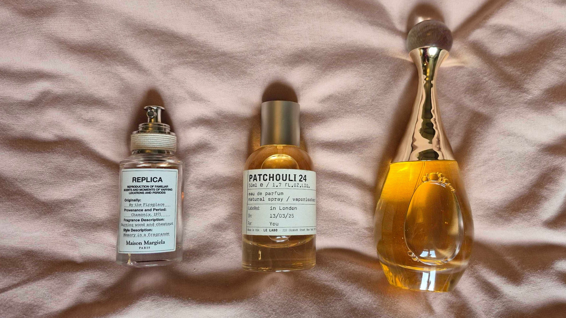 Bottles of the Maison Margiela&#039;s By The Fireplace perfume, Le Labo&#039;s Patchouli 24 and Dior&#039;s J&#039;adore l&#039;Or (all included in this guide), arranged and pictured on a pink bedding background