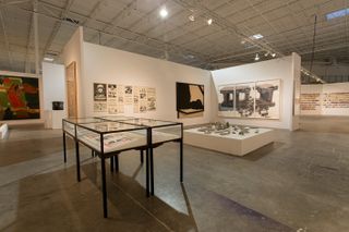 The exhibition space featuring a glass table display to the left, a series of posters on a wall and other artwork.