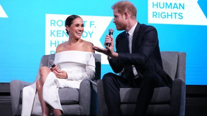 Prince Harry and Meghan Markle look at each other while speaking into microphones on a stage