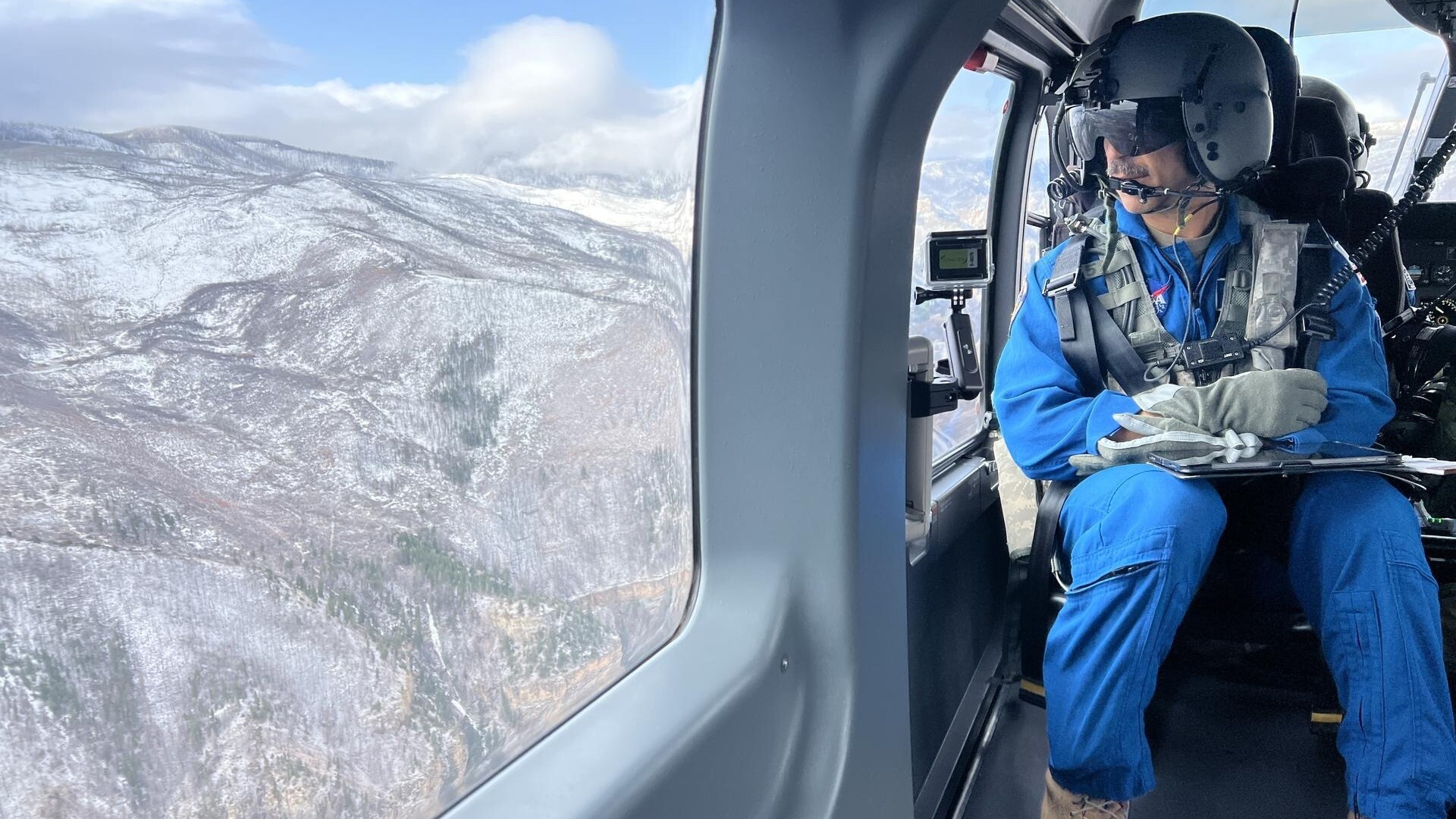 A man in a blue jumpsuit sits next to a plane window wearing a helmet and headset as he looks down over a series of snowy mountains