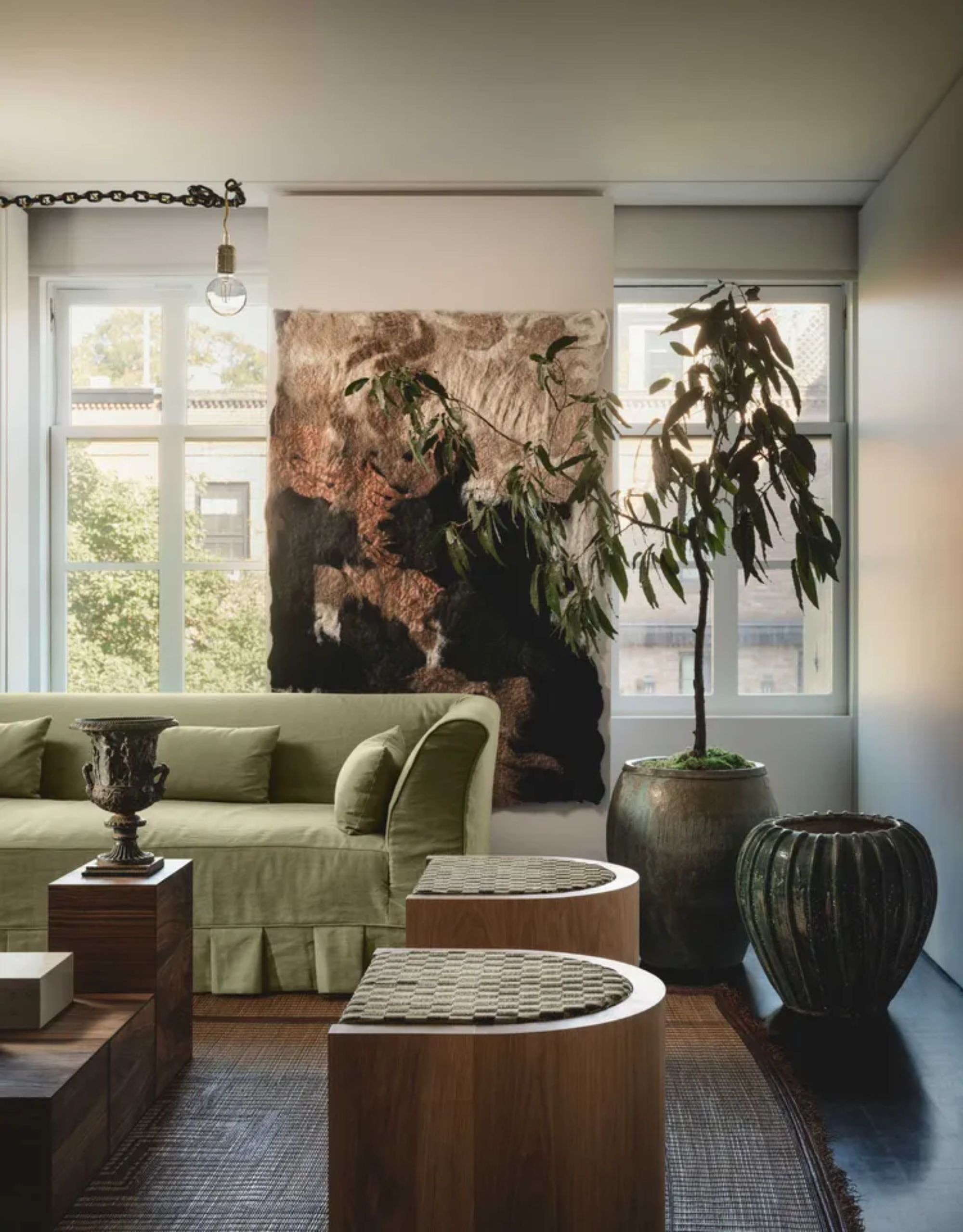 A white living room with a textured tapestry, a potted tree, a textured planter, a sofa with a green cover, a pair of stools, and a plinth with a goblet