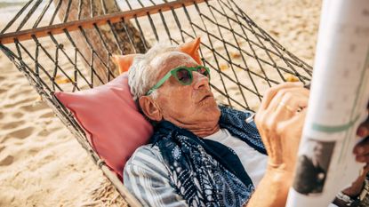 Senior man solving puzzle in the newspaper while lying down on a hammock at the beach