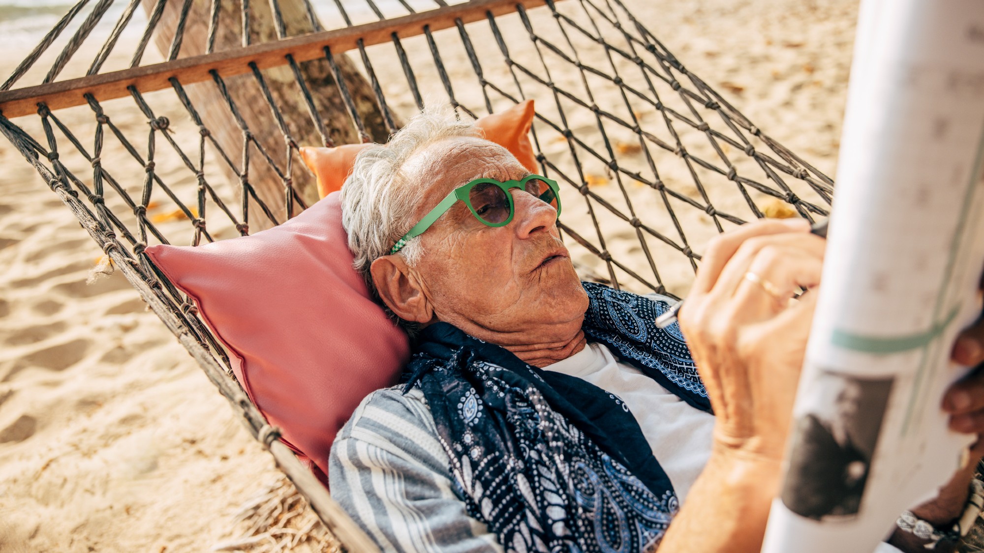 Senior man solving puzzle in the newspaper while lying down on a hammock at the beach