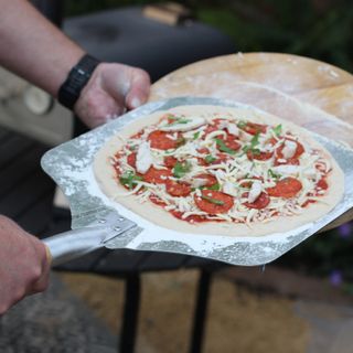 Uncooked pizza on a pizza paddle in front of a wood fired pizza oven
