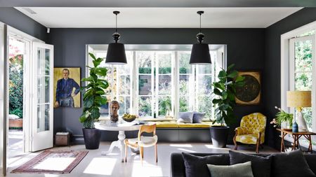 Image of a living room with lots of windows and natural light. The room is painted in a dark charcoal color and the ceiling is white. There are yellow accents in things like table lamps, bench seat cushions and accent chairs around the room.