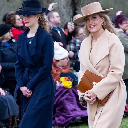 Sophie, the Duchess of Edinburgh wearing a tan coat and hat walking. next to Lady Louise Windsor in a navy coat with crowds of people behind them