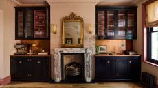 Veined marble fireplace in the middle of two built in kitchen cabinet unit. It's in a dark and moody utility room with dark academia feel, black cabinets, low lighting and gold and brass accents