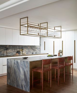 A white kitchen with a blue-green marble kitchen island, pink bar chairs, overhead glass storage, and hardwood floors