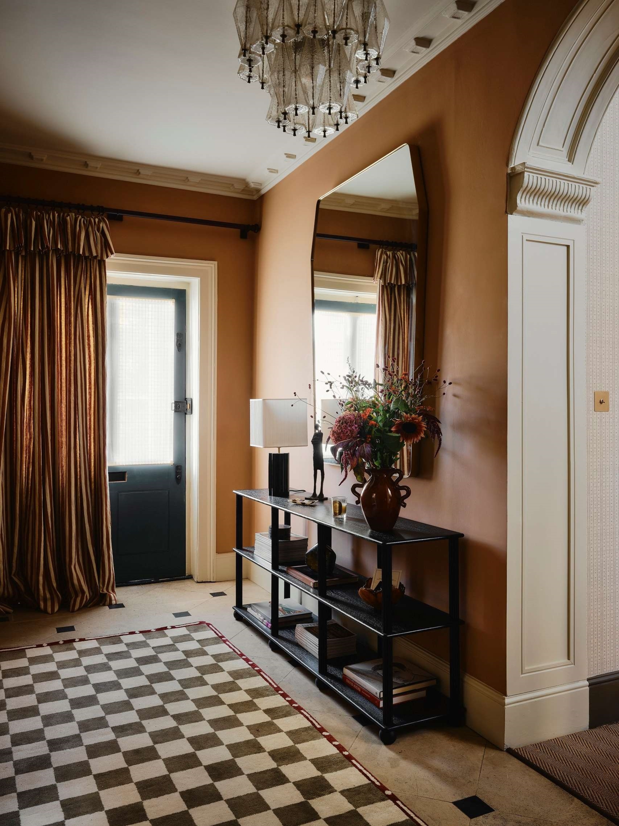 Image of a traditional entryway painted in a burnt orange color with off-white trim. There is a checkerboard rug on the ground and a striped curtain over the door. There is a black entry table with a mirror and a lamp and vase.