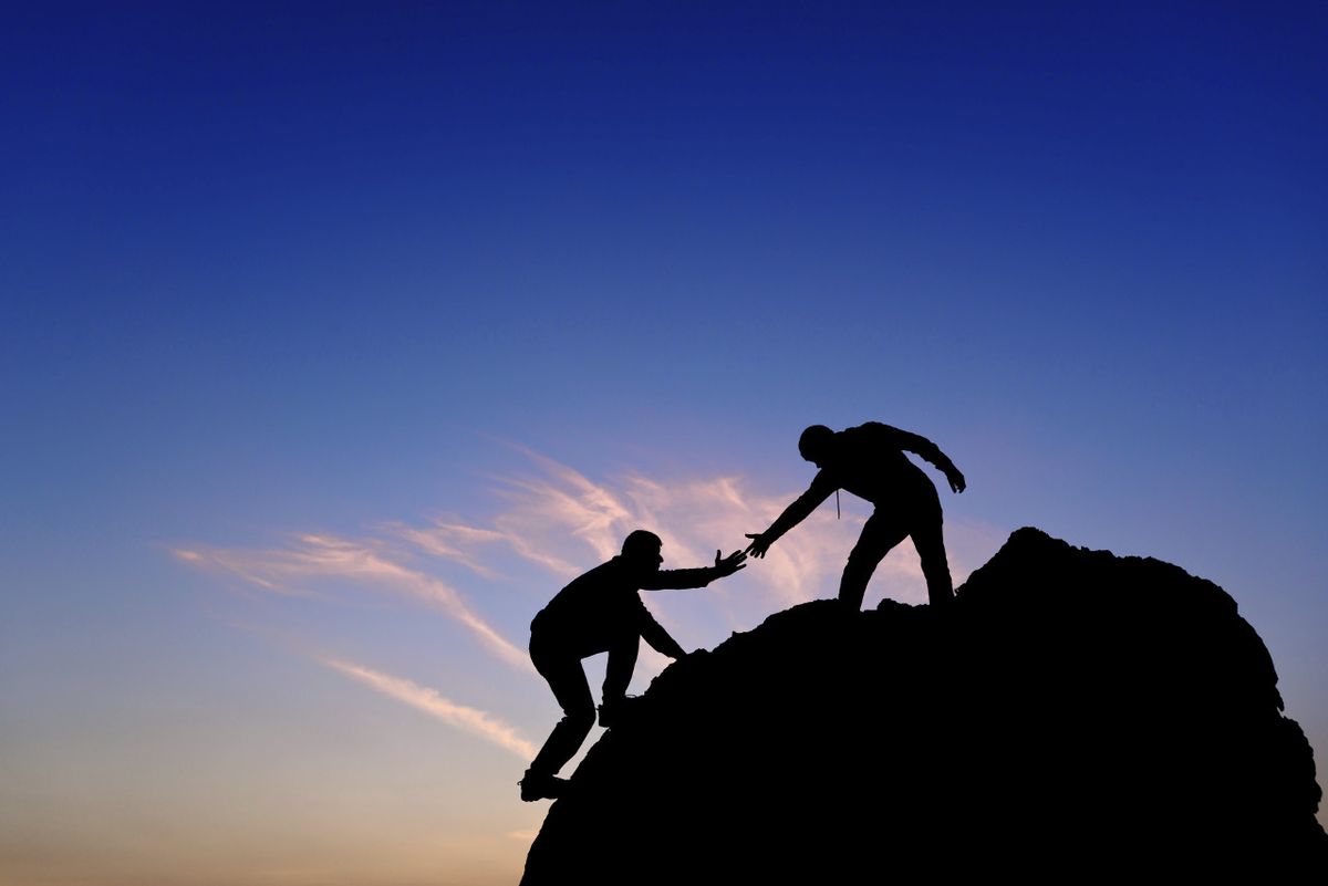 A figure in silhouette atop a mountain extends a hand to help another climber.