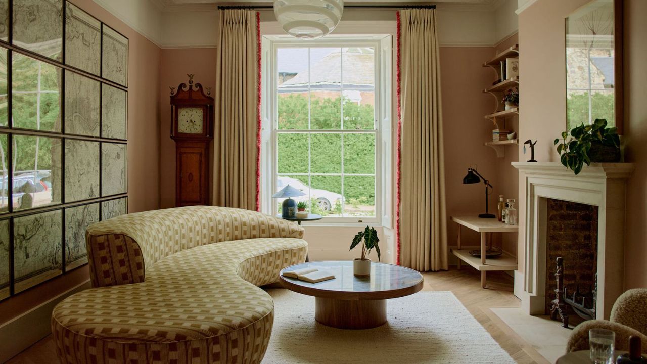 large soft pink painted living room with a floor to ceiling window and a large curved upholstered couch in front of a round coffee table and fireplace