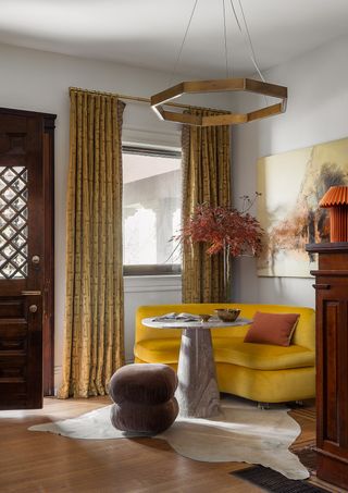 An entrance with a yellow couch, a metallic lighting piece above, an organic cream carpet, and long, printed curtains