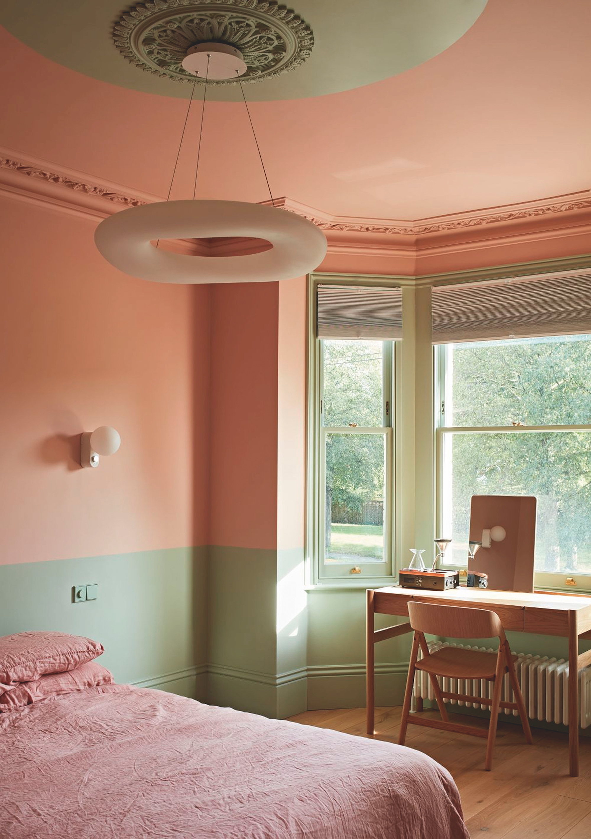 A peach and pistachio plastered bedroom with a large bay window to the right of the bed. There is detailed, traditional style cornicing and a modern chandelier above the bed.