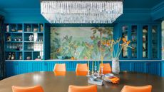 Large dining room with wooden oval eight seater in foreground with mid century style orange seats, with a floor-to-ceiling built in storage shelving and unit in the foreground. There is a tropical art wall in the background center, with large rectangular chandelier visible